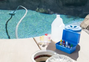 Pool maintenance equipment on a deck, including a vacuum hose in the water, test kit with colored solutions, a large white jug, and a blue container.