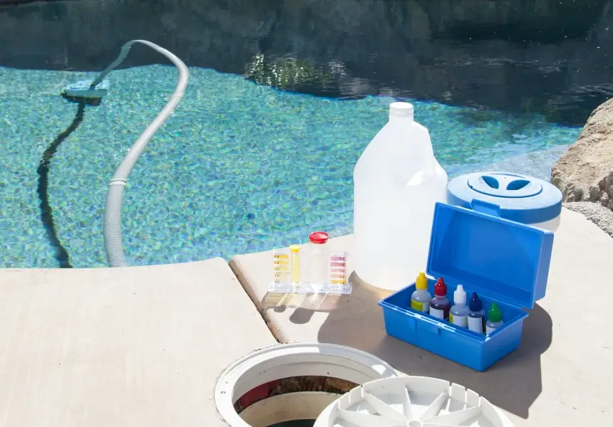 Pool maintenance equipment on a deck, including a vacuum hose in the water, test kit with colored solutions, a large white jug, and a blue container.