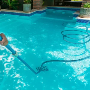 A person cleans a blue swimming pool with a vacuum hose and pole. Potted plants and a brick wall surround the pool area.
