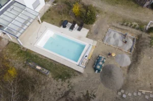 Aerial view of a rectangular swimming pool next to a house, surrounded by a garden. Nearby, three lounge chairs, a pile of gravel, and a small fenced gravel area are visible.