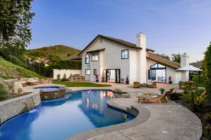 Two-story house with white exterior, large windows, and a sloped roof set by a curved pool and hot tub. Outdoor furniture is placed on a spacious patio surrounded by greenery and hills.