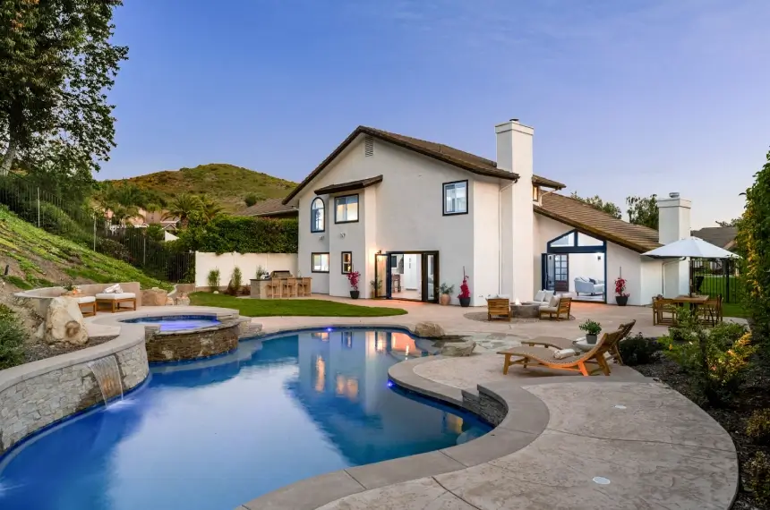Two-story house with white exterior, large windows, and a sloped roof set by a curved pool and hot tub. Outdoor furniture is placed on a spacious patio surrounded by greenery and hills.