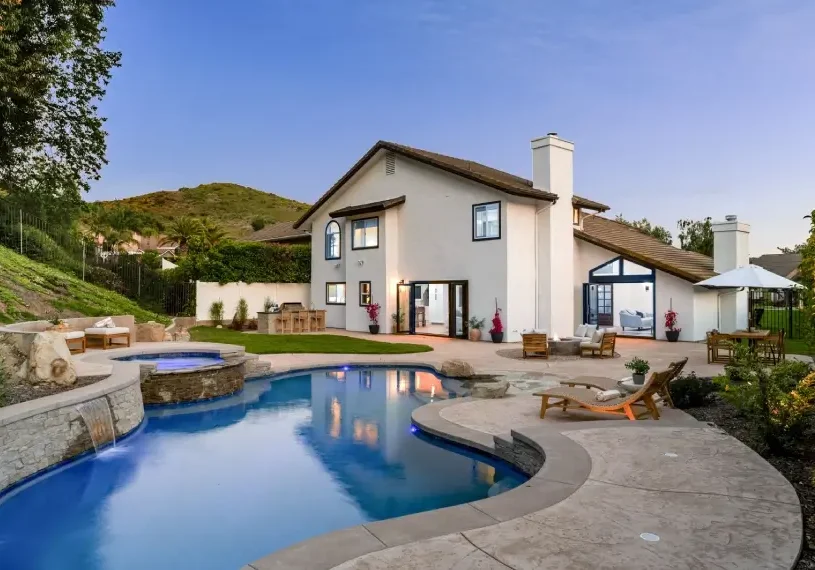 Two-story house with white exterior, large windows, and a sloped roof set by a curved pool and hot tub. Outdoor furniture is placed on a spacious patio surrounded by greenery and hills.